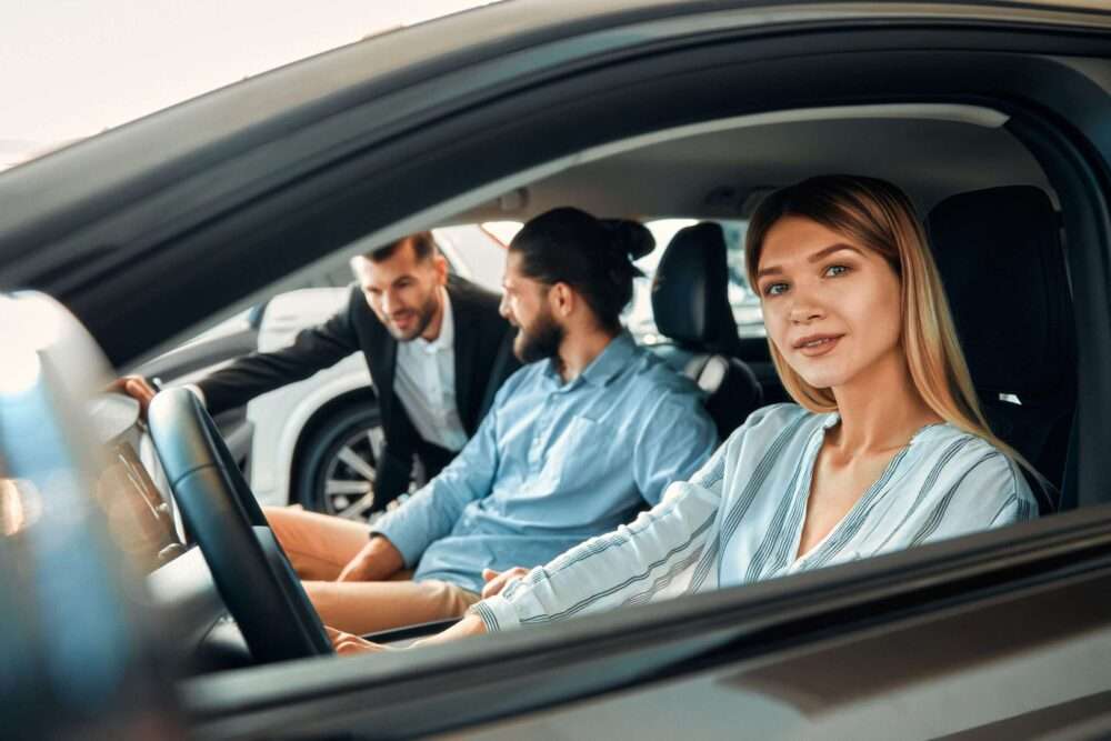 auto verkaufen - Eine lächelnde Frau sitzt auf dem Fahrersitz eines Autos, ein Mann auf dem Beifahrersitz. Ein weiterer Mann steht außerhalb des Autos, beugt sich vor und unterhält sich mit den beiden. Im Hintergrund sind Autos und ein Gebäude zu sehen. - Gebrauchtwagen Autohändler