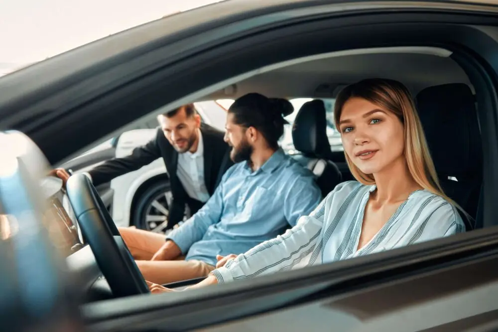 auto verkaufen - Eine lächelnde Frau sitzt auf dem Fahrersitz eines Autos, ein Mann auf dem Beifahrersitz. Ein weiterer Mann steht außerhalb des Autos, beugt sich vor und unterhält sich mit den beiden. Im Hintergrund sind Autos und ein Gebäude zu sehen. - Gebrauchtwagen Autohändler
