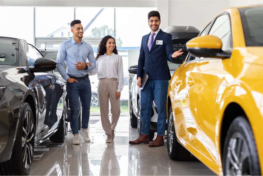 auto verkaufen - Ein Autoverkäufer zeigt einem lächelnden Paar in einem hellen Ausstellungsraum ein gelbes Auto. Das Paar geht gemeinsam, während der Verkäufer auf das Fahrzeug deutet. Weitere Autos sind im Ausstellungsraum zu sehen. - Gebrauchtwagen Autohändler