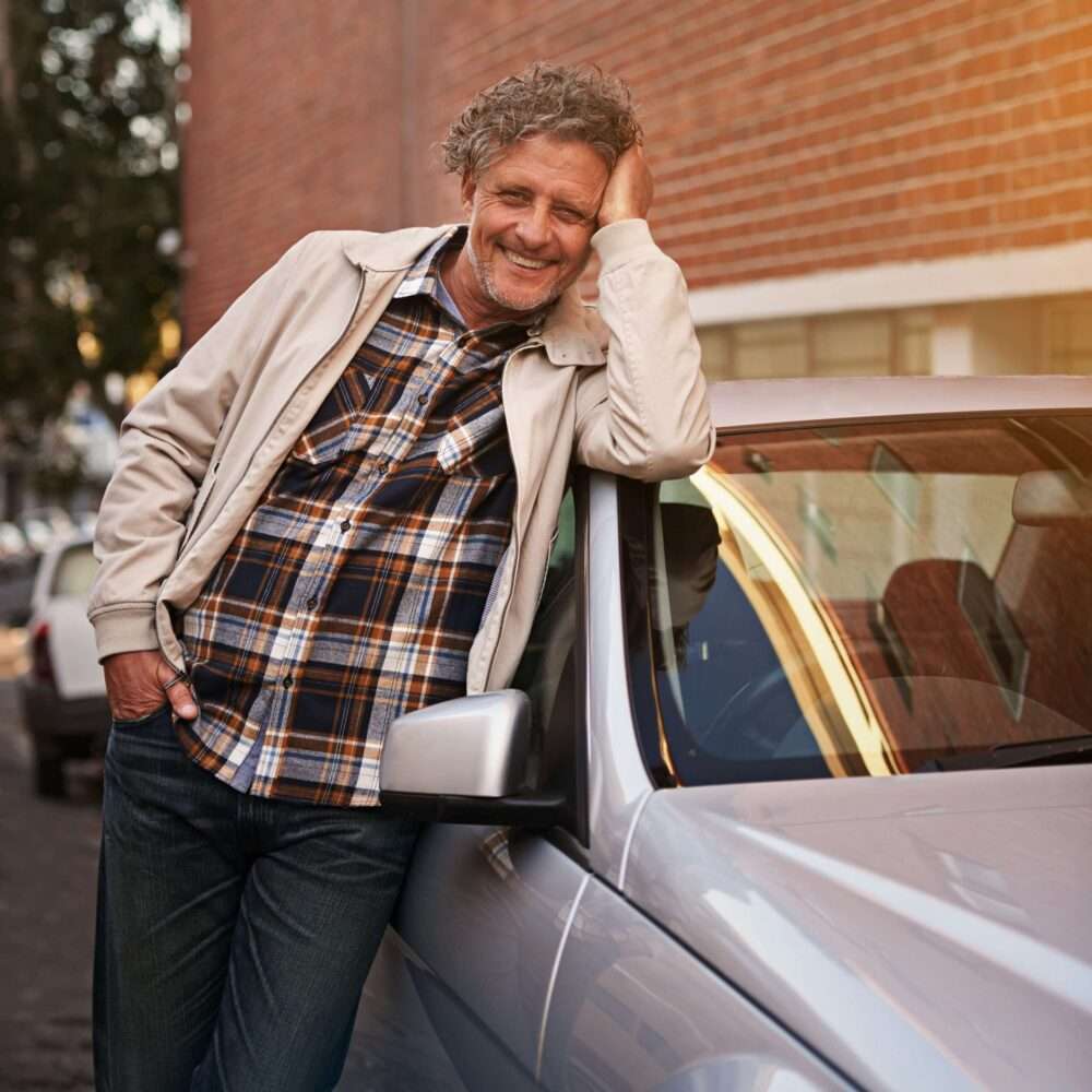 auto verkaufen - Ein Mann mittleren Alters mit lockigem grauem Haar und Bart lehnt lächelnd an einem silbernen Auto. Er trägt eine leichte Jacke über einem karierten Hemd und Jeans. Im Hintergrund sind eine Backsteinmauer und weitere parkende Autos zu sehen. - Gebrauchtwagen Autohändler